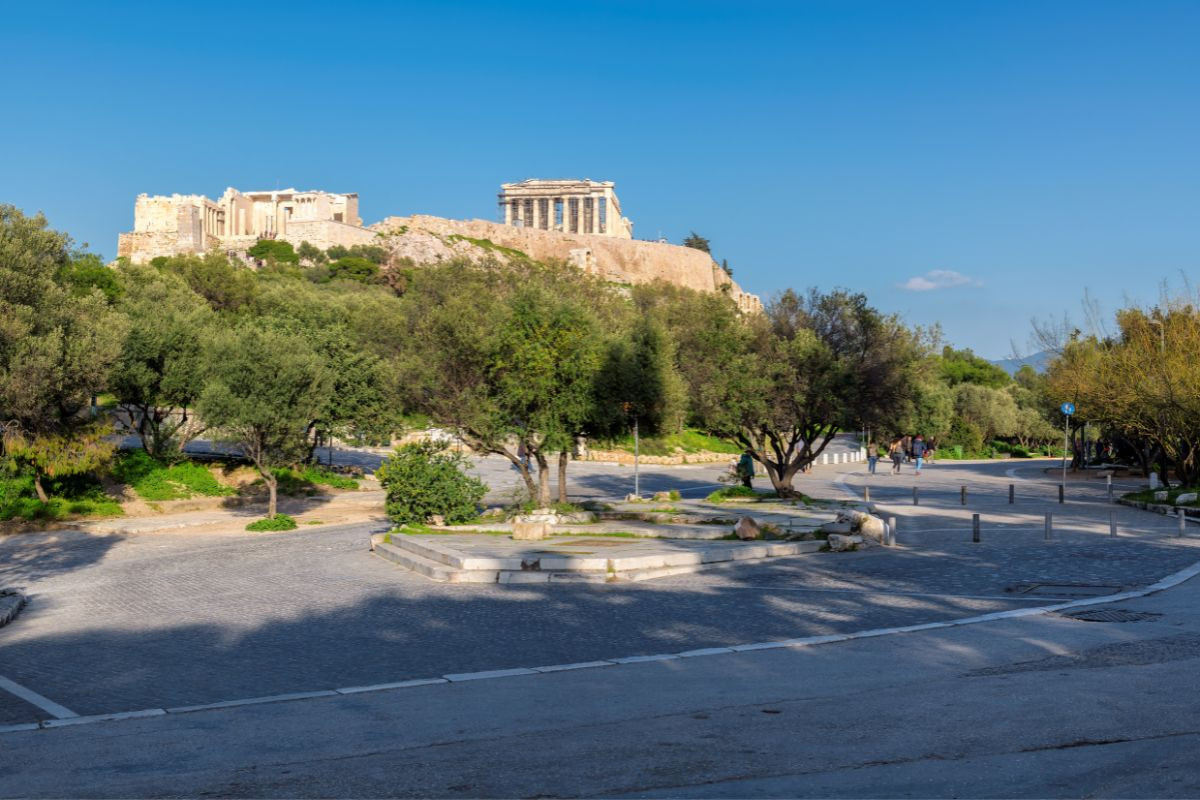 A paved area with trees leading up to the Acropolis in Athens, visible under a blue sky.